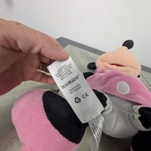 Puede incluir: Un mu&ntilde;eco de peluche de Minnie Mouse en rosa, negro y blanco. La mu&ntilde;eca tiene un vestido rosa, orejas negras y una cara blanca. Una etiqueta est&aacute; adjunta con texto, incluyendo "ALL NEW MATERIALS".