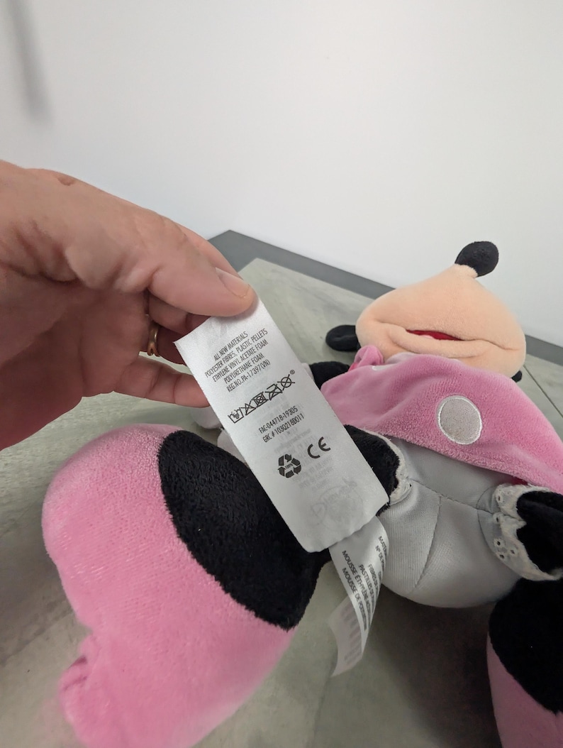 Puede incluir: Un mu&ntilde;eco de peluche de Minnie Mouse en rosa, negro y blanco. La mu&ntilde;eca tiene un vestido rosa, orejas negras y una cara blanca. Una etiqueta est&aacute; adjunta con texto, incluyendo "ALL NEW MATERIALS".