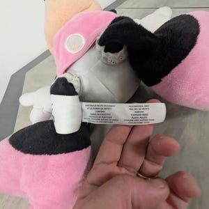 Puede incluir: Un peluche con un dise&ntilde;o rosa, negro y beige. El juguete tiene un lazo rosa, orejas negras y un estampado de lunares blancos y negros. La etiqueta del juguete enumera los materiales utilizados, incluidas las fibras de poli&eacute;ster y los gr&aacute;nulos de pl&aacute;stico.