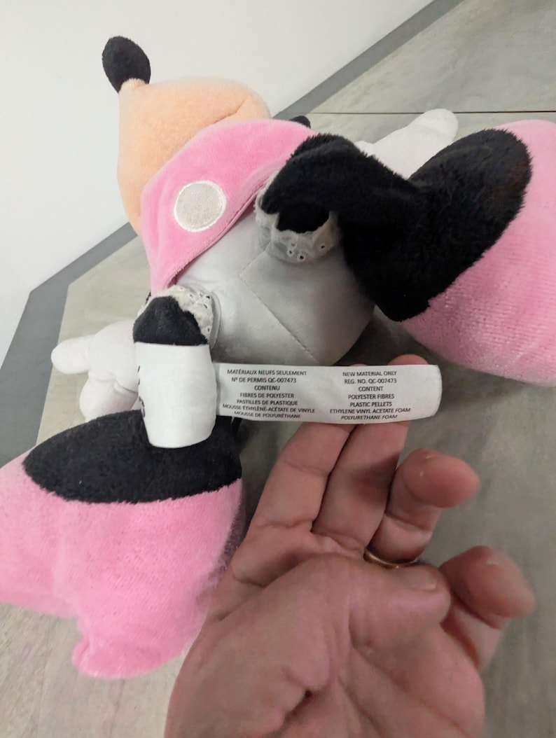 Puede incluir: Un peluche con un dise&ntilde;o rosa, negro y beige. El juguete tiene un lazo rosa, orejas negras y un estampado de lunares blancos y negros. La etiqueta del juguete enumera los materiales utilizados, incluidas las fibras de poli&eacute;ster y los gr&aacute;nulos de pl&aacute;stico.