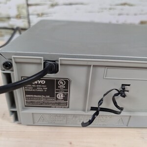 May include: A gray Sanyo VHR-H531 VCR with a black power cord plugged into the back. The back of the VCR has a UL Listed sticker and other printed information. The VCR sits on a light brown wooden surface.
