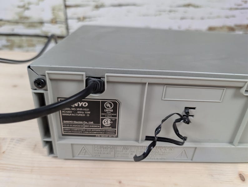 May include: A gray Sanyo VHR-H531 VCR with a black power cord plugged into the back. The back of the VCR has a UL Listed sticker and other printed information. The VCR sits on a light brown wooden surface.