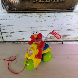 Puede incluir: Una moto de juguete amarilla y verde con Elmo montado. La figura roja de Elmo tiene un asiento azul y amarillo. Una bandera roja con "ELMO" está adjunta. Una cuerda roja está unida a la moto.