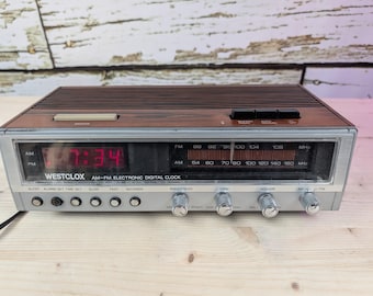 May include: A vintage Westclox AM-FM electronic digital clock with a wood-grain top and silver face. The red digital display shows the time as 7:34. Features include radio tuning, volume, and alarm controls. The clock is a retro style.