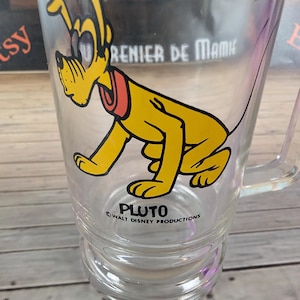 May include: Clear glass mug featuring Pluto, a yellow cartoon dog with a red collar, from Walt Disney Productions. The mug has a handle and the word "PLUTO" is printed on the front.