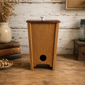 May include: A wooden container with a dark brown top and a white knob. The container is made of light brown wood and has a circular hole near the bottom. It is sitting on a wooden surface.