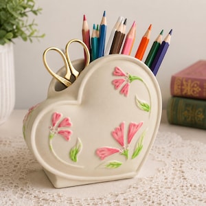 Peut inclure: Un organisateur de bureau en céramique crème en forme de cœur avec des motifs floraux roses en relief et des feuilles vertes. L'organisateur contient une paire de ciseaux dorés et une collection de crayons multicolores dans des tons de bleu, rouge et marron.