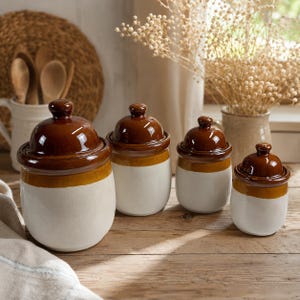 Peut inclure: Quatre pots de cuisine en céramique avec couvercles. Les pots sont blancs avec une bande marron et jaune et un couvercle marron brillant. Ils sont disposés sur une surface en bois, avec un panier tressé et des ustensiles en arrière-plan.