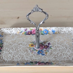 Peut inclure: Plateau de service rectangulaire à deux niveaux avec une poignée argentée. Le plateau présente un motif floral doré avec des fleurs roses, violettes et bleues. La poignée est de conception décorative et ouverte.