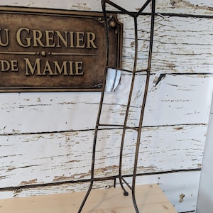 May include: A tall, wrought-iron display stand with a weathered, dark brown finish. The stand has a rectangular frame with multiple horizontal bars and a three-legged base. The background features a wooden wall and a sign that reads "U Grenier de Mamie."
