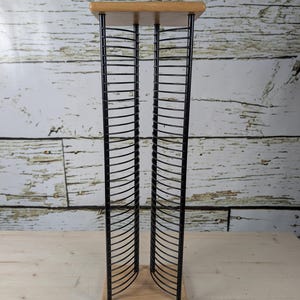 Peut inclure: Un présentoir à CD en métal noir avec une base et un dessus en bois. Le présentoir est composé de séparateurs verticaux en fil de fer incurvés pour ranger les CD. Les éléments en bois sont de couleur claire et naturelle, contrastant avec le métal noir.