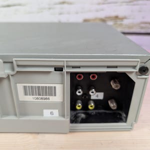 May include: A gray electronic device with a series of input/output ports. The ports include red, white, and yellow connectors, along with coaxial cable inputs. A barcode label and the number "6" are visible.