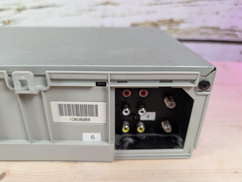 May include: A gray electronic device with a series of input/output ports. The ports include red, white, and yellow connectors, along with coaxial cable inputs. A barcode label and the number "6" are visible.