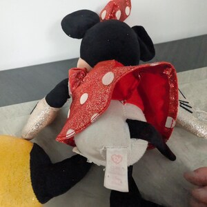 May include: A Minnie Mouse plush toy, viewed from the back, features a red dress with white polka dots and a matching bow. The toy has black ears, a black tail, and yellow shoes. The arms are silver glitter. A tag is visible.