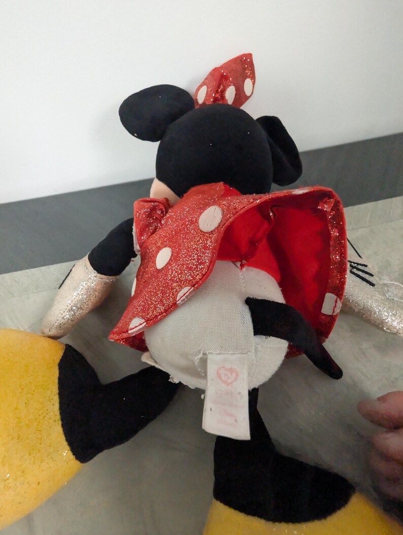 May include: A Minnie Mouse plush toy, viewed from the back, features a red dress with white polka dots and a matching bow. The toy has black ears, a black tail, and yellow shoes. The arms are silver glitter. A tag is visible.