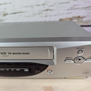 May include: Silver VHS player with a black front panel. The player has buttons for pause, rewind, and play. The text "4 HEAD Hi-Fi VCR 19-MICRON HEADS" is printed on the front.