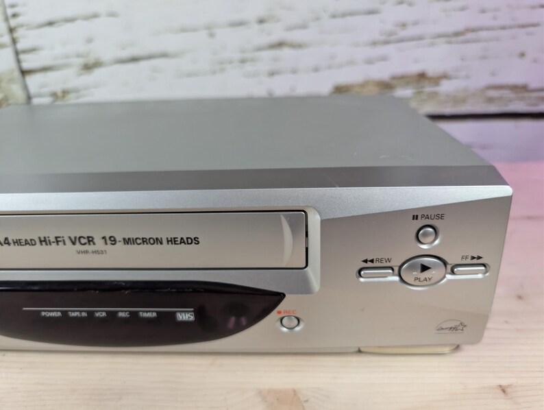 May include: Silver VHS player with a black front panel. The player has buttons for pause, rewind, and play. The text "4 HEAD Hi-Fi VCR 19-MICRON HEADS" is printed on the front.