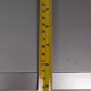 May include: A yellow and black measuring tape is positioned vertically against a gray surface. The tape shows measurements in inches, with clear markings and numbers. The image highlights the tape's precision and readability.
