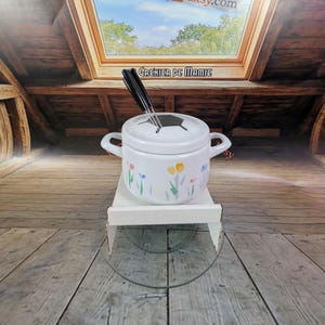 May include: A white ceramic fondue pot with a lid and handles, decorated with colorful floral designs. Two black fondue forks are inserted into the lid. The pot sits on a white shelf and a glass surface.