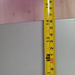 May include: A yellow and black measuring tape is positioned next to a gray rectangular object. The tape shows measurements in inches, with the object's height at approximately 15 inches.