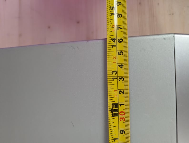 May include: A yellow and black measuring tape is positioned next to a gray rectangular object. The tape shows measurements in inches, with the object's height at approximately 15 inches.