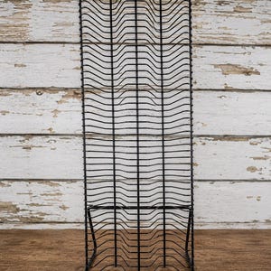 Peut inclure: Un grand support de rangement vertical en fil métallique noir. La structure présente une série de séparateurs horizontaux ondulés disposés en grille, soutenus par une base solide et surmontés de cinq petits embouts arrondis.