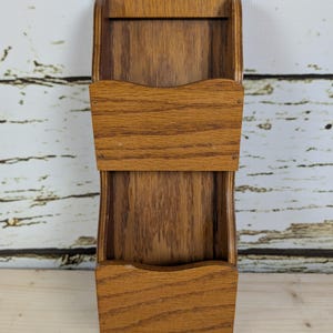 May include: A vintage wooden wall-mounted letter holder with two compartments. The holder is made of light brown wood with a natural grain pattern. The top has a curved design, and the compartments have a scalloped edge.