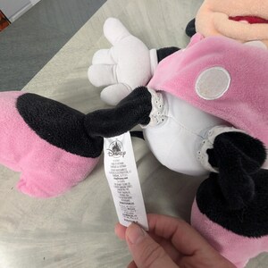 Puede incluir: Un peluche de Minnie Mouse, con un vestido rosa con lunares blancos, brazos y piernas negros y guantes blancos. El peluche tiene una boca roja y una etiqueta con el logo de Disney.