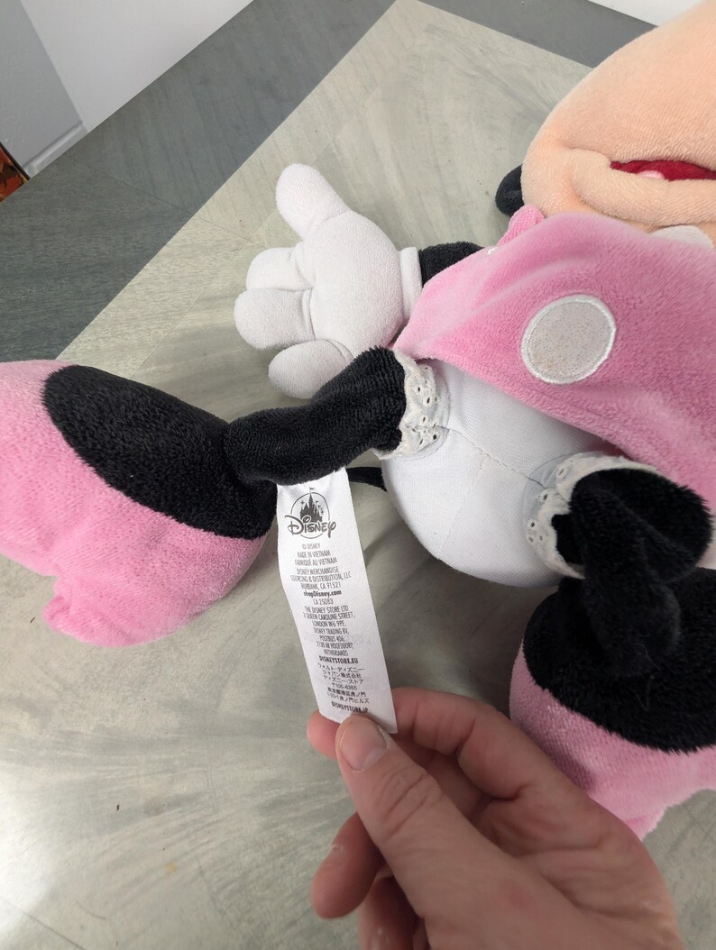Puede incluir: Un peluche de Minnie Mouse, con un vestido rosa con lunares blancos, brazos y piernas negros y guantes blancos. El peluche tiene una boca roja y una etiqueta con el logo de Disney.