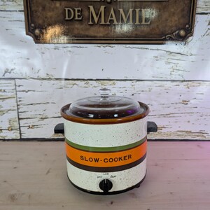 Peut inclure: Un autocuiseur vintage avec un extérieur crème moucheté, avec des rayures orange et vertes. Les mots "SLOW-COOKER" sont imprimés sur le devant. Il a un couvercle en verre et un bouton de commande noir avec les réglages "OFF", "LOW" et "HIGH".