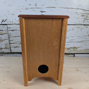 Può includere: Un contenitore in legno con coperchio e pomello bianco. Il contenitore è realizzato in legno marrone chiaro con la parte superiore marrone scuro. Ha un foro circolare sul davanti ed è posto su una superficie di legno chiaro.