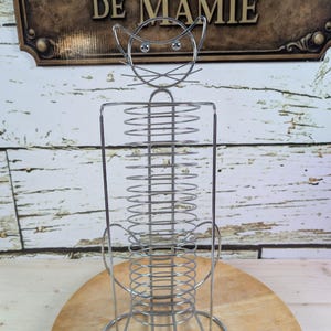 Może przedstawiać: Chromowany metalowy serwetnik w kształcie kota. Projekt obejmuje kocią twarz na górze, prostokątny korpus ze spiralą z drutu i zaokrągloną podstawą. Uchwyt znajduje się na drewnianej powierzchni.