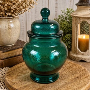Peut inclure: Un bocal en verre de couleur teal avec un couvercle, présentant une surface texturée. Le bocal a un corps arrondi et un petit bouton rond sur le dessus du couvercle. Le bocal est posé sur un plateau en bois.
