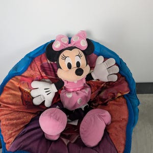 Puede incluir: Una mu&ntilde;eca de peluche de Minnie Mouse con un vestido rosa con lunares blancos y un lazo a juego. La mu&ntilde;eca tiene orejas negras y una cara sonriente. Est&aacute; sentada sobre una manta colorida estampada con un borde azul.