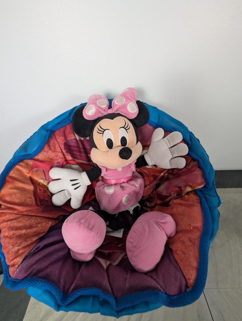 Puede incluir: Una mu&ntilde;eca de peluche de Minnie Mouse con un vestido rosa con lunares blancos y un lazo a juego. La mu&ntilde;eca tiene orejas negras y una cara sonriente. Est&aacute; sentada sobre una manta colorida estampada con un borde azul.