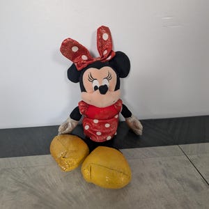 May include: A Minnie Mouse plush toy with a red dress with white polka dots, a red bow with white polka dots, and yellow glittery shoes. The toy has black ears and a black face with a smiling expression.