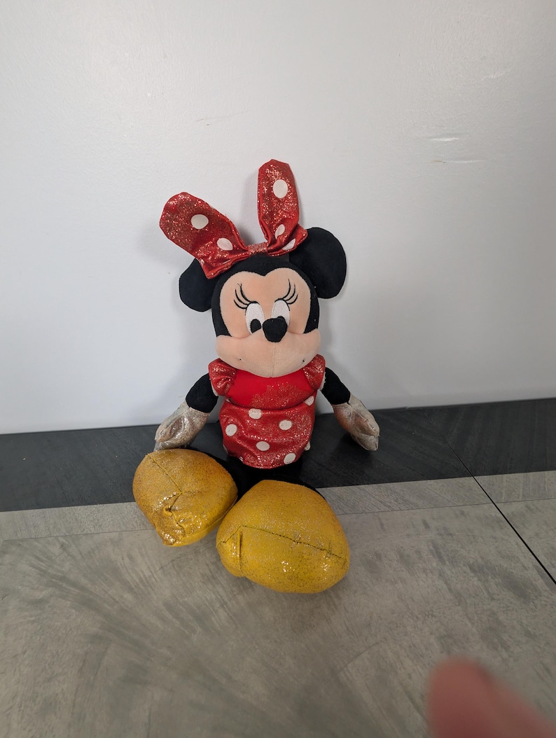 May include: A Minnie Mouse plush toy with a red dress with white polka dots, a red bow with white polka dots, and yellow glittery shoes. The toy has black ears and a black face with a smiling expression.