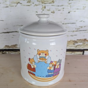 May include: White ceramic cookie jar with a lid. The jar features a colourful illustration of cartoon cats, one holding a plate of biscuits. The cats are dressed in various outfits. The jar has a handle on the lid.