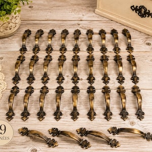 Könnte beinhalten: Eine Sammlung von 29 antiken bronzefarbenen Schrankgriffen. Jeder Griff hat ein geschwungenes Design mit floralen Verzierungen. Die Griffe sind auf einer hellen Holzoberfläche angeordnet. Der Text "29 POIGNÉES" ist sichtbar.