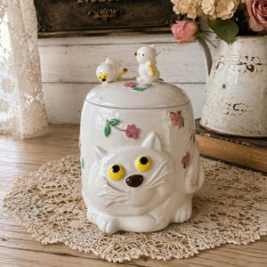 Peut inclure: Un pot en céramique blanc avec un motif de visage de chat, des yeux jaunes et un nez brun. Le couvercle présente deux petites figurines d'oiseaux et des motifs floraux. Le pot est décoré de fleurs roses et de feuilles vertes.