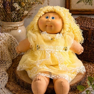 Puede incluir: Una muñeca Cabbage Patch Kid vintage con cabello rubio rizado y ojos azules. La muñeca lleva un vestido amarillo con ribete de encaje blanco y está sentada sobre una rodaja de madera. La piel de la muñeca es de color melocotón.