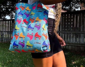 May include: A blue tote bag featuring a repeating pattern of colorful roller skates in shades of red, yellow, purple, and orange. The bag is carried by a person, and the background includes a wooden fence and green grass.
