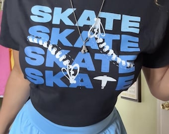 Könnte beinhalten: Schwarzes T-Shirt mit dem Wort "SKATE" in Blau, viermal wiederholt mit einem stilisierten Skate-Design. Das Shirt wird mit hellblauen Sporthosen kombiniert. Eine silberne Halskette mit einem Skate-Anhänger ist ebenfalls sichtbar.