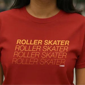 Puede incluir: Una camiseta roja con la frase "ROLLER SKATER" repetida en un patrón amarillo-naranja. La camiseta tiene cuello redondo y mangas cortas. Las letras "HSD" están impresas en la parte inferior.