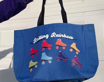 May include: A blue tote bag with black handles featuring the text "Rolling Rainbow" in white script. The bag displays a colorful array of roller skates in red, orange, yellow, green, blue, purple, and pink.