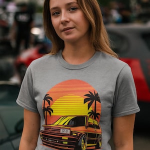 May include: Grey t-shirt with a retro car graphic set against a sunset and palm trees. The car is orange and yellow, with black detailing. The sunset is a blend of orange and yellow.