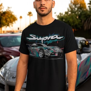 May include: Black t-shirt featuring a graphic of a Supra sports car in shades of gray, teal, and pink. The word "Supra" is displayed above the car in a stylized font. The shirt is worn by a person.