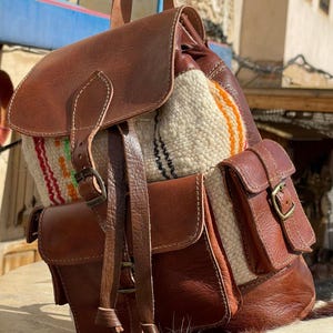 Pode incluir: Uma mochila de couro marrom com fecho de aba e dois bolsos laterais. O painel central é feito de um tecido tecido creme com listras coloridas. A bolsa tem alças ajustáveis e uma alça superior.