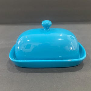 May include: A bright turquoise ceramic butter dish with a lid. The dish is rectangular with rounded corners and a glossy finish. The lid has a small, round knob on top. The butter dish is designed to hold a standard size of butter.