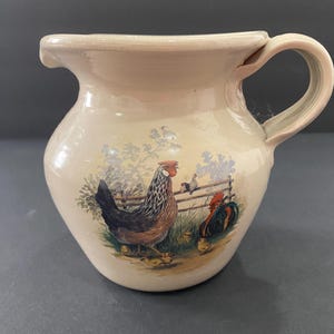 May include: A cream-colored ceramic pitcher featuring a painted scene of a hen with chicks and a rooster near a wooden fence. The pitcher has a rounded body, a wide spout, and a curved handle on the side, with a smooth, glossy glazed finish.